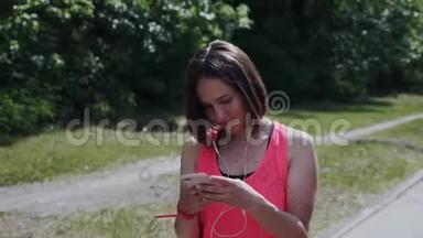 戴着耳机的可爱女孩穿过公园。 迷人的年轻女士戴着耳机在发短信。 看智能手机的女孩